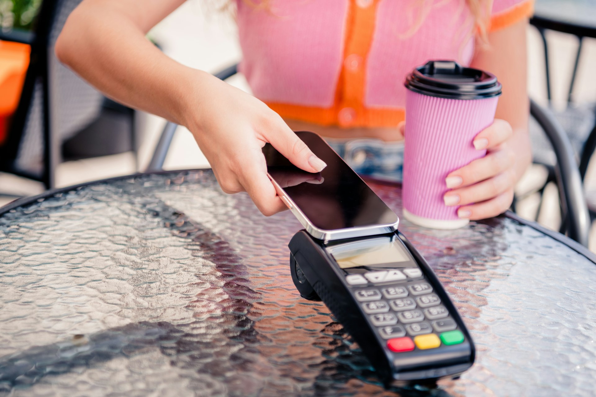 Frau sitzt an Tisch mit Kaffebecher und bezahlt mit Handy an Kartenterminal - kontaktloses Zahlen